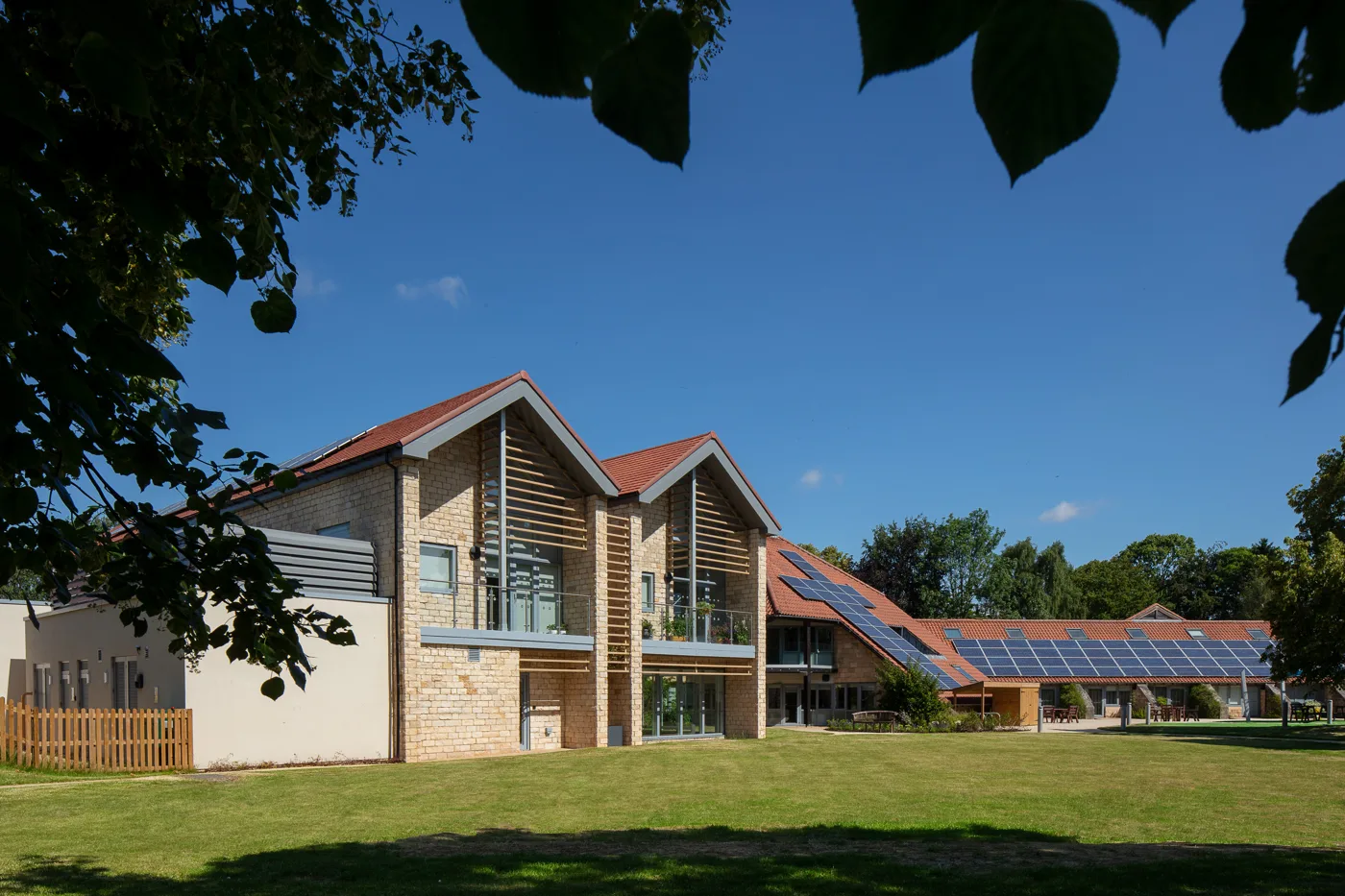 A two-storey stone building with gabled roofs, timber slats and balconies, beside a lawn, a path and a bench, framed by tree leaves under a blue sky
