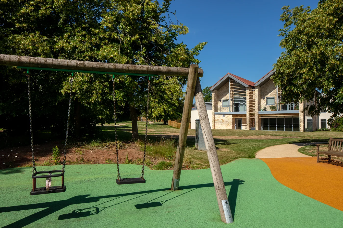 A wooden swing frame with two swings on green play surfacing, with a lawn, trees and a modern stone building behind