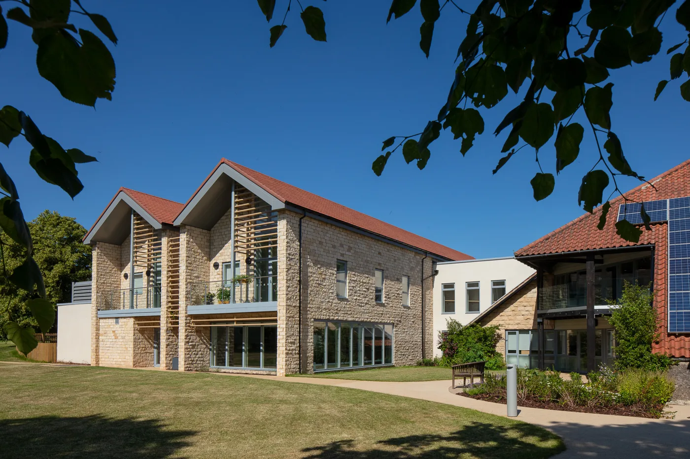 A long stone and render building with red tiled roofs and rows of solar panels, beside a wide lawn and trees