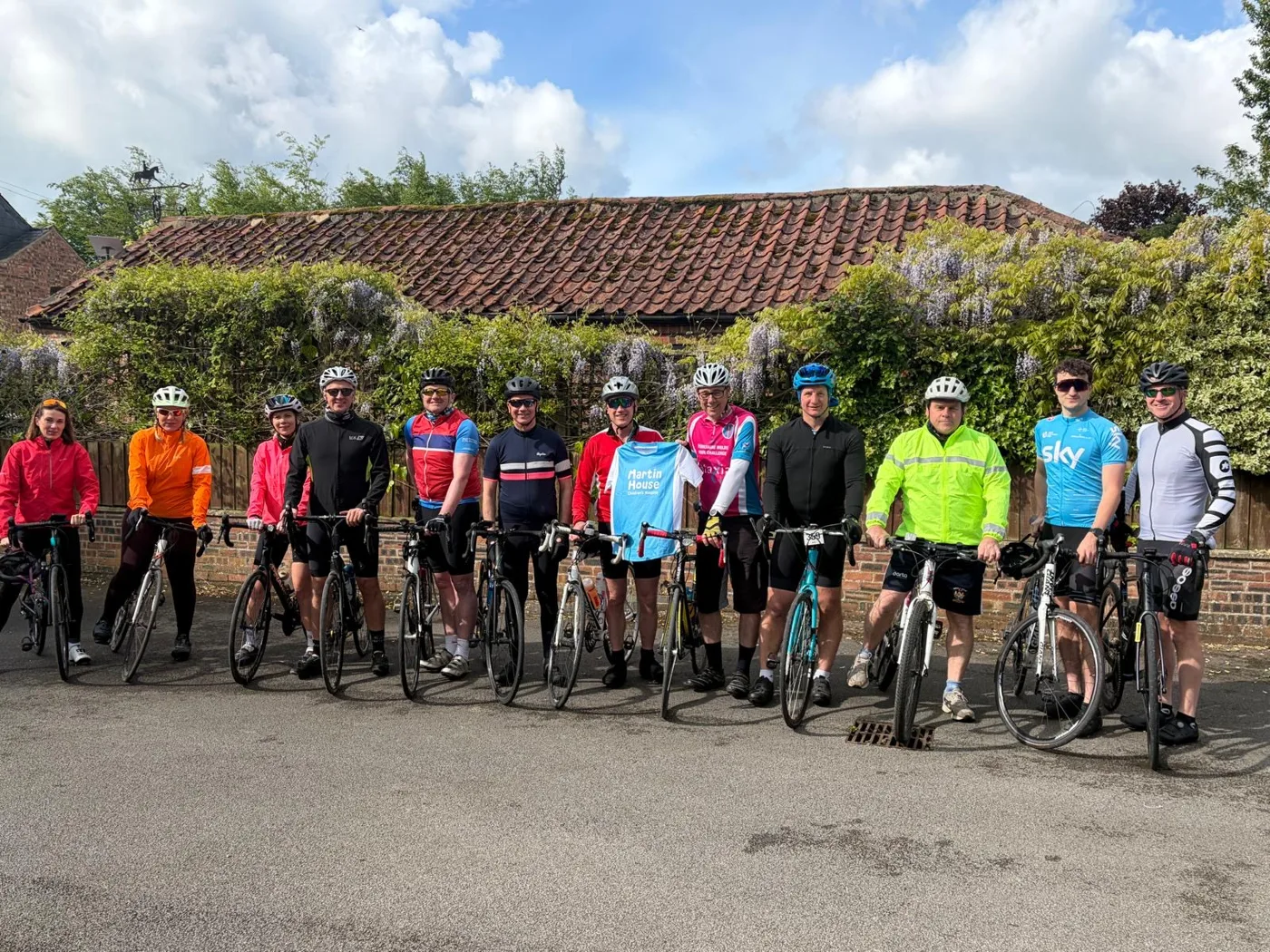 About a dozen cyclists in helmets and bright cycling jerseys lined up with their road bikes in a lane beside a wisteria-covered wall